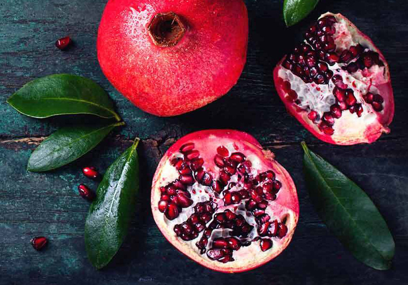 Two halves of a pomegranate beside a whole pomegranate. Leaves and seeds surround the fruit.