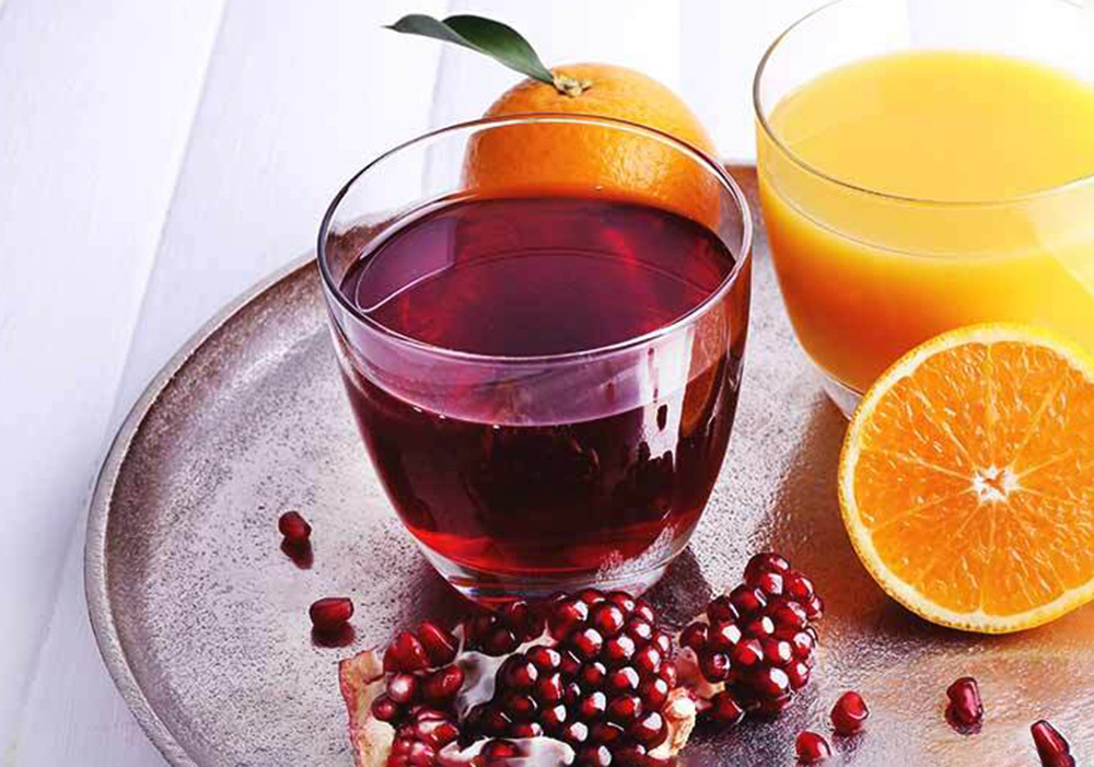 Glass of red pomegranate juice beside glass of orange juice. Pomegranate seeds and oranges in foreground and background.